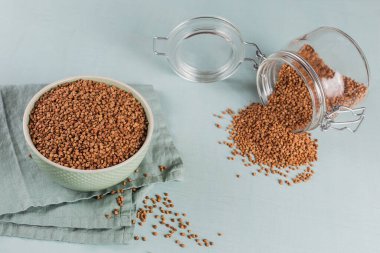 Pişmemiş organik karabuğday taneleri bir kasede ve açık mavi arka planda cam kavanozda. Sağlıklı ve diyet gıda konsepti.