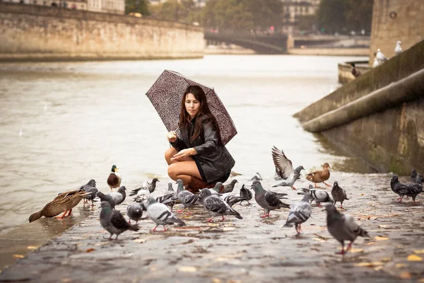 A pretty woman with an umbrella feeds birds on the embankment of the river
