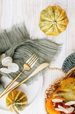 Top view of vertical autumn table with green napkin, gold cutlery, white plate and decorative gourds on rustic wood surface