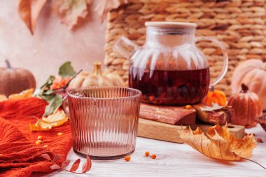 Bitkisel deniz dikenli çaydanlığın içinde, yapraklar, balkabakları ve kırmızı bir eşarpla süslenmiş kırsal bir sonbahar masasında boş bir cam bardağın yanında..