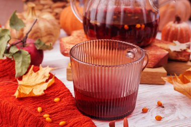 Sonbahar yaprakları, kırmızı elma ve rahat bir ortamda balkabakları ile çevrili bitkisel çayla dolu kaburgalı cam bardak.