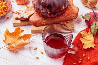 Kırsal beyaz ahşap masa üzerinde kırmızı bitkisel çaylı cam bardak. Sonbahar yapraklı, elmalı, deniz dikenli ve çaydanlıklı.