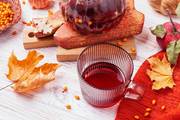 Kırsal beyaz ahşap masa üzerinde kırmızı bitkisel çaylı cam bardak. Sonbahar yapraklı, elmalı, deniz dikenli ve çaydanlıklı.