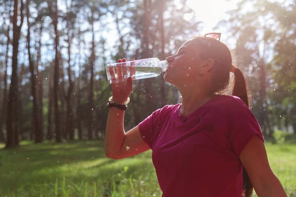 Ormanda güneş ışınları ve su damlacıklarıyla su içen olgun bir kadın.