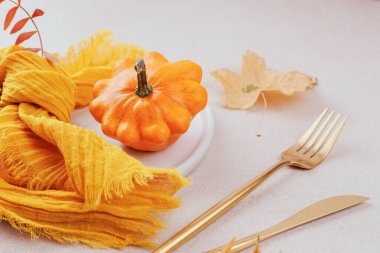 Sonbahar dekoru portakal kabağı, sarı eşarp peçetesi, altın çatal bıçak takımı ve kurumuş akçaağaç yapraklarıyla yakın plan.