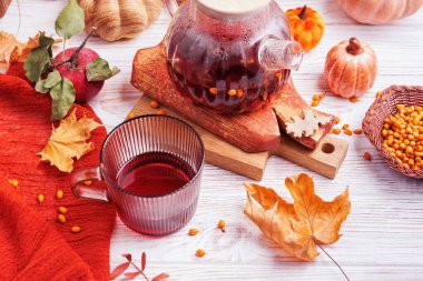 Düz cam bardak ve kırmızı bitkisel çayla çevrili çaydanlık. Beyaz ahşap üzerinde balkabakları, elmalar, yapraklar ve böğürtlenler.