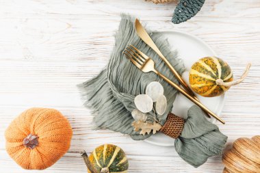 Yeşil peçete, altın çatal bıçak, turuncu iplik kabağı ve beyaz ahşap arka planda çizgili su kabağı olan düz bir sonbahar masa dekoru.