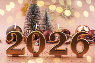 Altın 2026 mum, Noel ağacı süslemeleri kar taneleri ve yumuşak pembe şenlik arka planında parıltılı mücevherlerle dimdik ayakta duruyor.