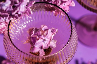 Yumuşak mor tonlarla çevrili leylak çiçekleri ve altın köpüklü şarapla kaplı desenli bir bardağın yakın görüntüsü