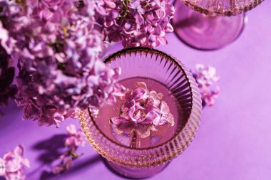 Çiçekler ve buketlerle çevrili kristal bir bardağın içinde yüzen leylak çiçekli pembe bir içeceğin yukarıdan görünüşü