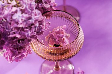 Pembe bir yüzey üzerinde leylak çiçekleriyle çevrili, içinde yüzen leylak çiçekli, ışıl ışıl bir şarap kadehi.