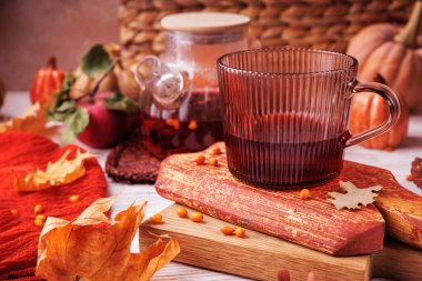 Kırsal ahşap yüzeyde sonbahar yaprakları ve elma yapraklarıyla kırmızı bitki çayı ve çaydanlık ile tırtıklı cam bardak.