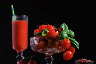 Cam bir kasede fesleğenli olgun kiraz domatesleri ve kırmızı yüzeyde uzun bir bardakta domates suyu.