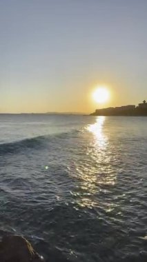 Altın bir günbatımının altında, sudaki yansımalarla ve sakin sahil atmosferiyle yumuşak okyanus dalgaları yuvarlanıyor.