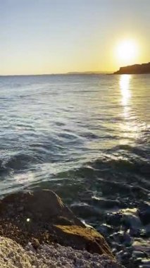 Okyanus dalgaları, kayalık kıyı şeridinde altın gün batımında parıldıyor. Güneş, suyu ve huzurlu kıyı manzarasını yansıtıyor.