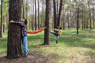 Anne hamak ipini eforla çekerken, genç çocuk çam ormanında ipini sabit tutuyor.