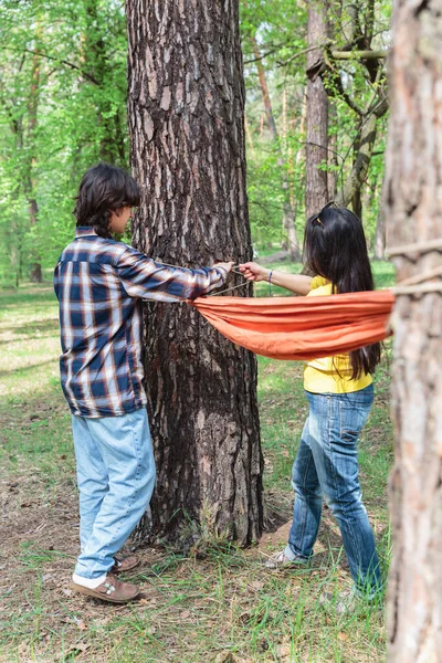 Genç çocuk ve anne ormanda kamp yaparken senkronize bir şekilde hamak ipi çekiyorlar.