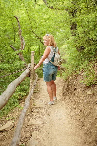 Orta yaşlı şort giymiş bir kadın sırt çantasıyla orman yolunda yürüyor. Çekici kadın gezgin yürüyüş parkurunda rahatlıyor ve doğanın tadını çıkarıyor. Seyahat ve sağlıklı yaşam tarzı
