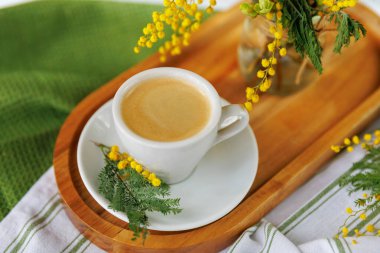 Taze sarı mimoza çiçekleri ve yumuşak bir masa örtüsüyle süslenmiş, ahşap tepside bir fincan kahveyle sıcacık bir bahar sabahı düzeni..