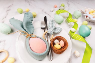 Paskalya tabağı setine yakın kumaş tavşan peçetesinde pembe yumurta, yumurtalı yuva, kurdele ve bahar çatal bıçak takımı.