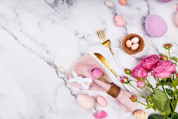 Paskalya temalı masa dekorunun üst görüntüsü pembe güller, pastel yumurtalar, altın kaplar ve tavşan peçetesi halkası.