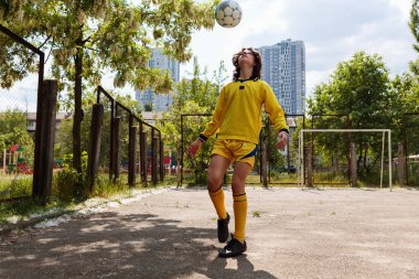 Genç futbolcu çocuk açık hava eğitimi sırasında şehir binaları ve yaz yeşilliği ile oyun alanında top çeviriyor.