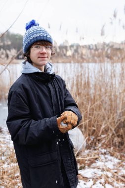 Kış kıyafetleri içinde genç bir çocuk, elinde yumuşak bir ifadeyle kartopu tutuyor, gölün kenarında duruyor.
