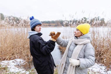 Genç bir çocuk, göl kenarındaki soğuk hava yürüyüşü sırasında sıcak eldiven giymesi için annesinden yardım alır.