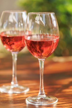 Ahşap bir masada, yeşil bahçe arka planında, gül şarabıyla dolu iki bardak.