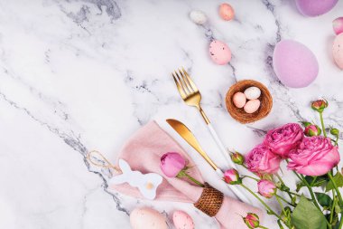 Peçete, çatal bıçak, güller, benekli yumurtalar ve mermer yüzeyde şekerleme kovanlarıyla zarif Paskalya kompozisyonu.