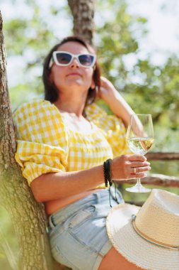Ağacın yanında oturan ve elinde beyaz şarap bardağı tutan rahat olgun bir kadın. Parlak yaz ormanlarında, kırsal yaşam tarzında ve özgürlük kavramında.