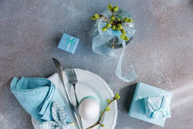 Beyaz yumurta, mavi peçete, hediye kutuları, çatal bıçak takımı ve tomurcuklu vazo.