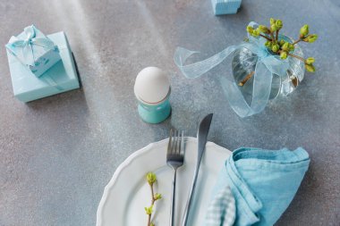Paskalya temalı yumurta, hediye kutuları, çiçek açan vazo ve pastel yüzeyde beyaz tabak.