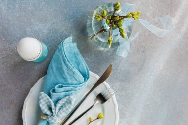 Tabakta yumurta, mavi peçete, çatal bıçak ve çiçek vazosu olan Paskalya bezi.