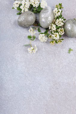 Benekli paskalya yumurtaları, narin beyaz çiçek çiçekleri ve yeşil yapraklarla desenli gri arka planda düzenlenmiş ve bahar tatili konseptini kopyalayan.