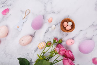 Pastel Paskalya yumurtaları, yuvada şeker yumurtaları, pembe güller ve mermer arka planda beyaz keçeli tavşan süsü.