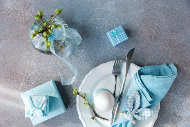 Tepesinde beyaz yumurta, kurdele vazosu, mavi hediye kutuları ve çatal bıçak takımı olan Paskalya düzeneği.