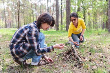 Genç bir çocuk ve annesi yeşil ormandaki açık hava aktiviteleri sırasında birlikte kamp ateşi yakmaya odaklanıyor.