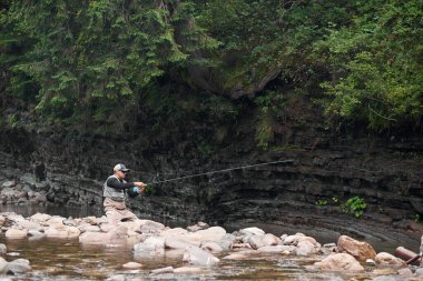 Dağ nehrinde üniformalı ve şapkalı genç bir adam balık tutuyor.