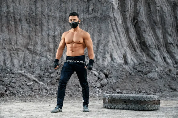 Bodybuilder in mask using tyre for training at sand pit