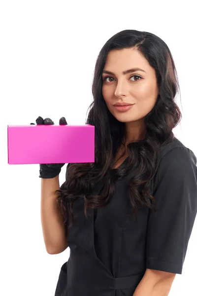 Привабливий косметолог в уніформі тримає рожеву паперову коробку — стокове фото
