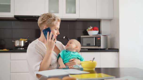 Kvinna pratar på mobilen medan du sitter i köket med barnet — Stockvideo