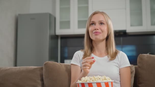 Joven rubia viendo películas y comiendo palomitas de maíz en el sofá — Vídeos de Stock