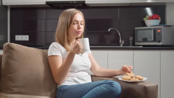 Ontspannen vrouw die koekjes eet en thee drinkt op de bank — Stockvideo