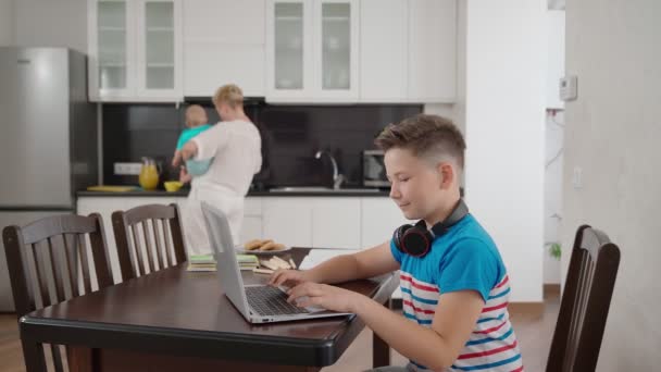 Chłopiec za pomocą laptopa podczas gotowania matki w kuchni — Wideo stockowe
