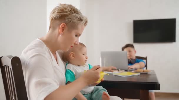 Matka łyżka karmienie dziecko chłopiec podczas nastolatek za pomocą laptopa — Wideo stockowe