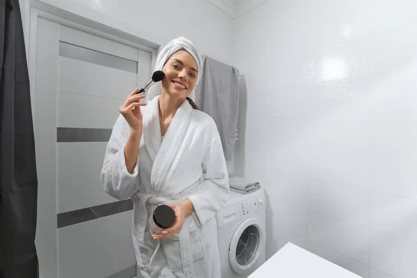 Mujer joven sonriente en bata haciendo maquillaje en casa. —  Fotos de Stock