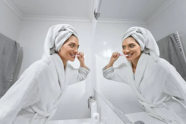 Woman in white robe with towel on head brushing teeth. — Stock Photo, Image