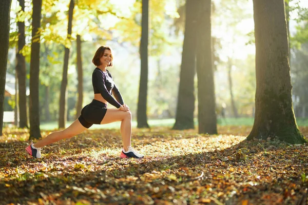 Uśmiechnięta kobieta w stroju aktywnym rozciągająca nogi w parku — Zdjęcie stockowe