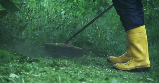 Man in gele rubberen laarzen gras maaien met elektrische maaier — Stockvideo